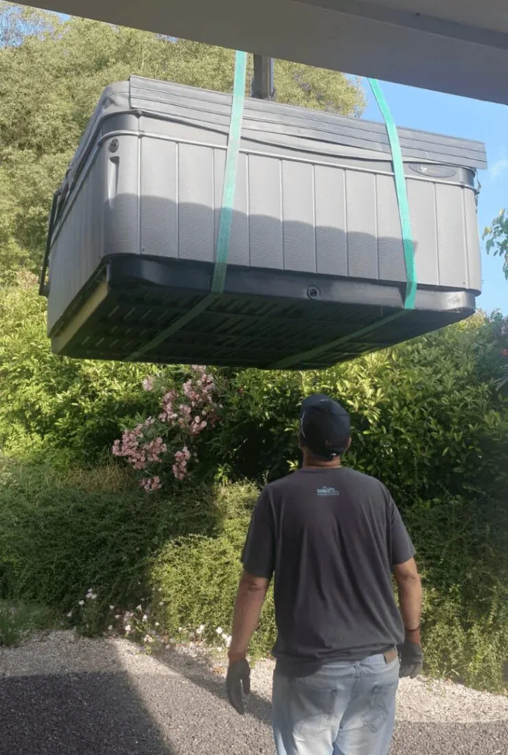 Un homme marche sous un spa suspendu aux sangles de la grue, dans un jardin arboré