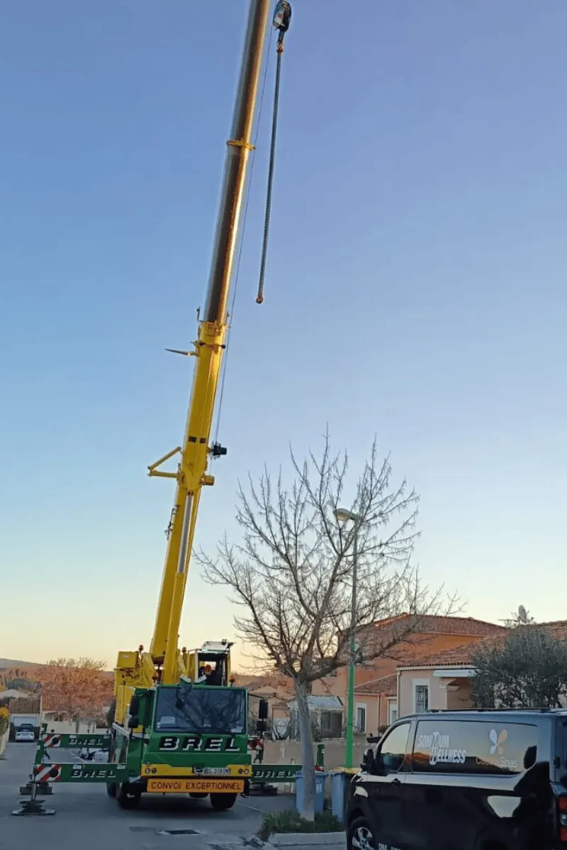 Grue mobile jaune des Transports Brel dépliée dans une rue de Mèze, fourgon Sommum Wellness garé devant
