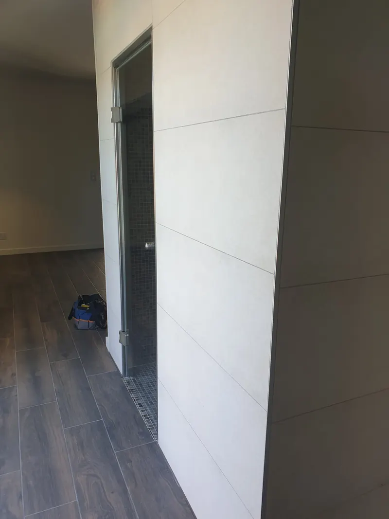 Cabine de hammam habillée de grands carreaux clairs, porte vitrée à cadre inox, dans une pièce à Perpignan