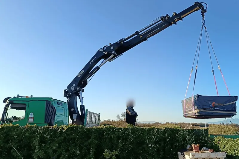 Camion-grue des Transports Brel soulevant le spa bâché au-dessus de la haie, à Leucate