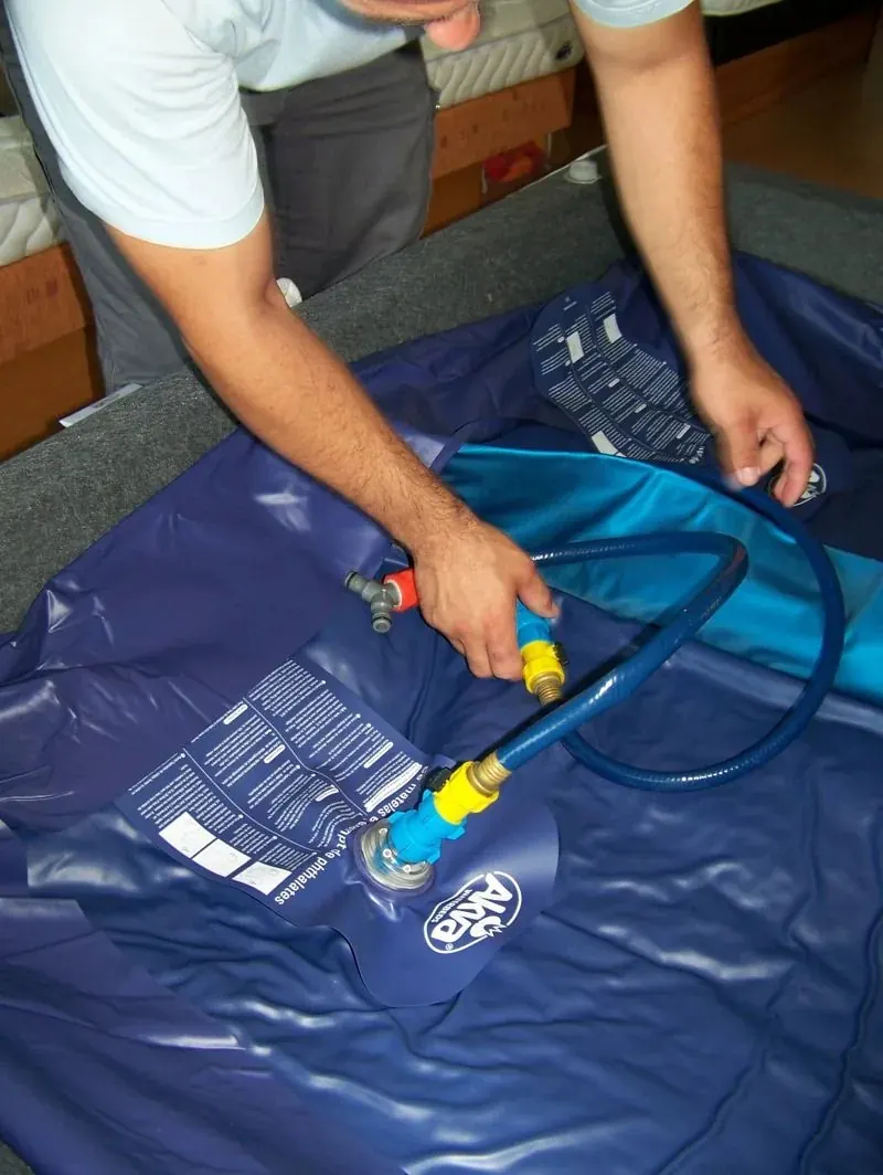 Deux mains raccordent le tuyau de remplissage à la valve d'un matelas de lit à eau, dans son liner bleu