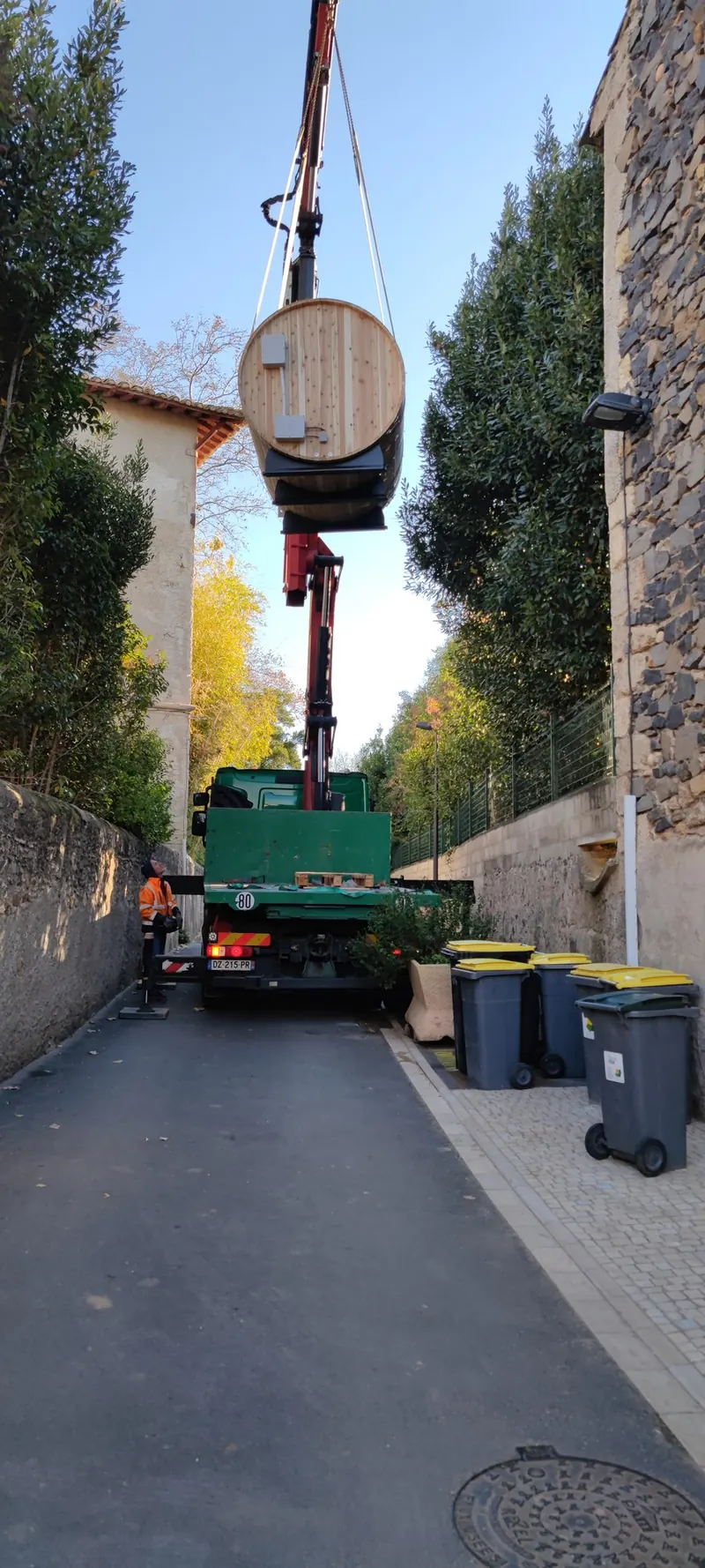Sauna tonneau en bois suspendu à la grue, camion et ruelle étroite entre deux murs