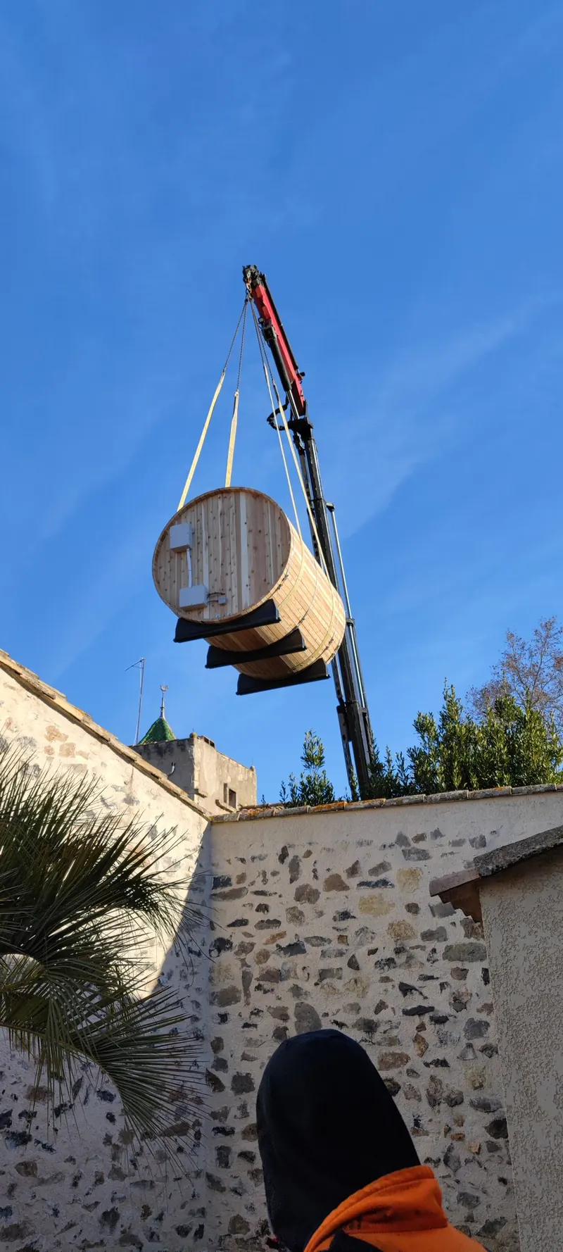 Sauna tonneau suspendu au-dessus d'un mur de pierre, palmier et flèche de la grue