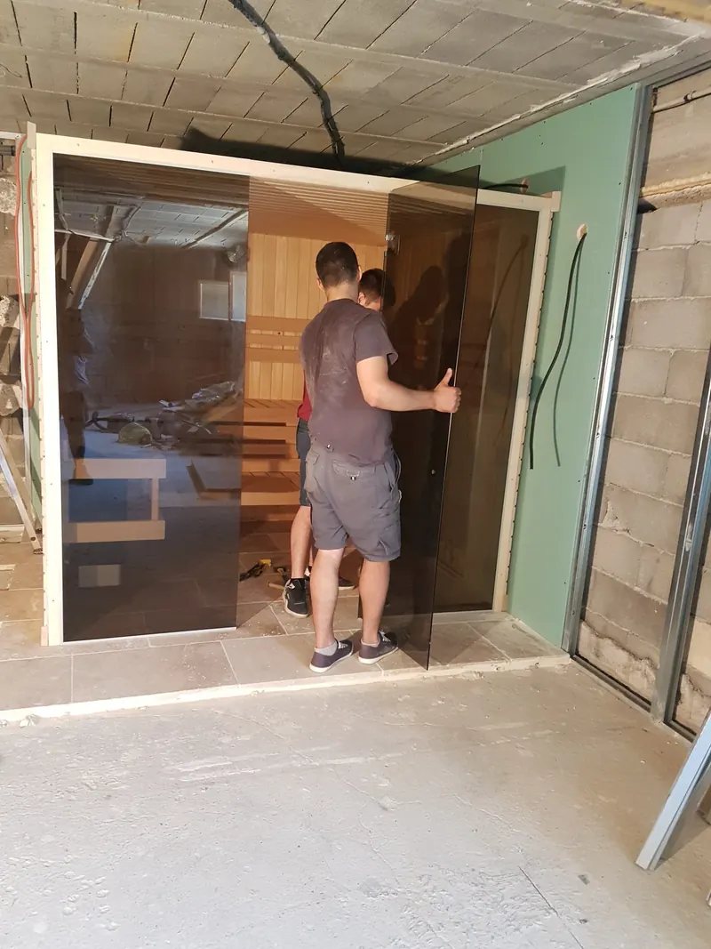 Deux installateurs de dos mettent en place la porte en verre fumé du sauna à Mèze