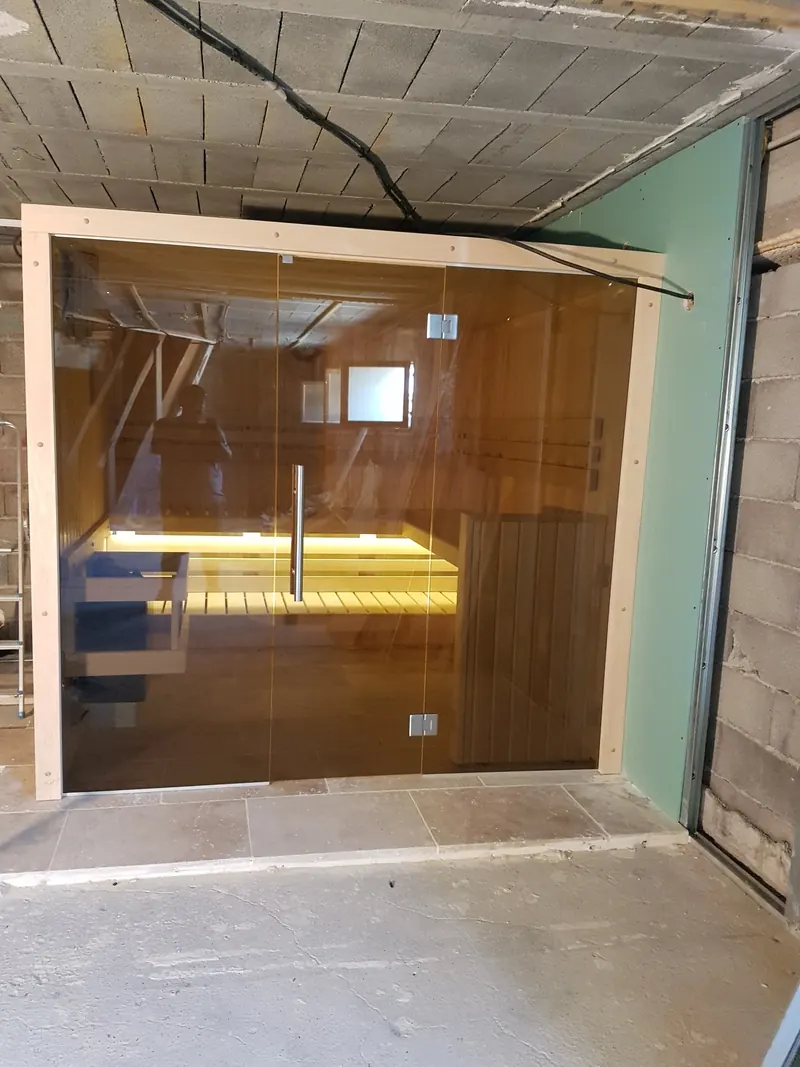 Sauna terminé avec façade en verre fumé et banquettes éclairées, dans une pièce en travaux à Mèze
