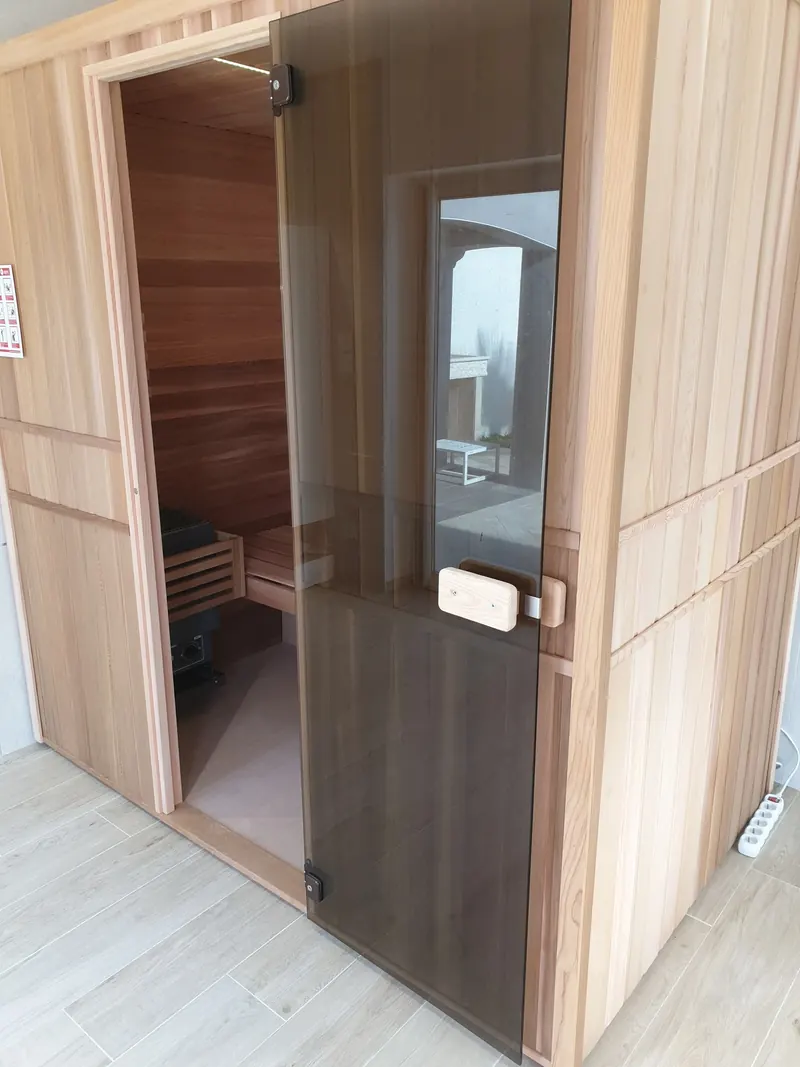 Sauna en bois clair à porte en verre fumé, poêle à pierres visible à l'intérieur, à Vic-la-Gardiole