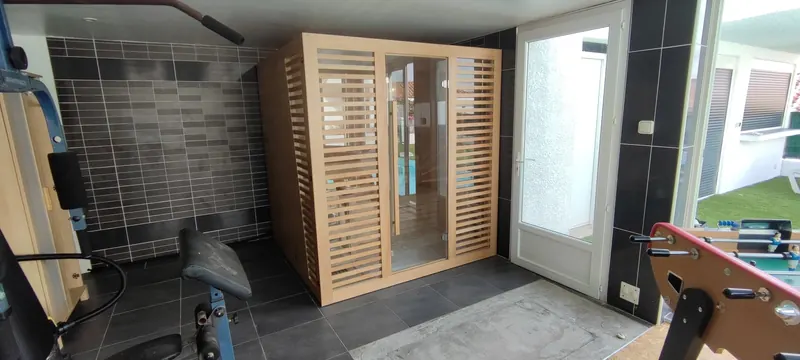 Sauna en bois clair, porte à lames fines, dans une salle carrelée, vue d'ensemble