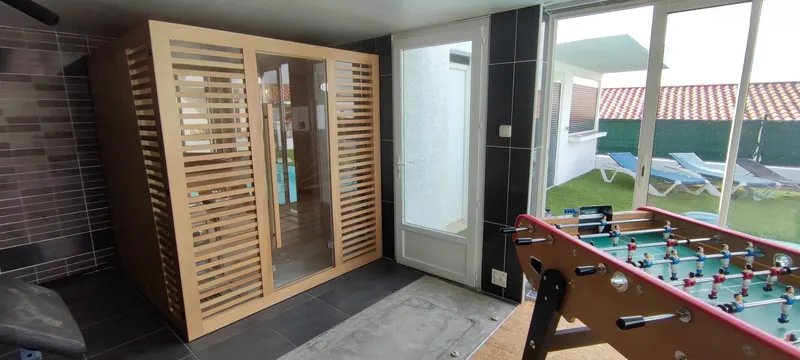 Sauna dans la salle carrelée grise, baies vitrées ouvrant sur la piscine turquoise