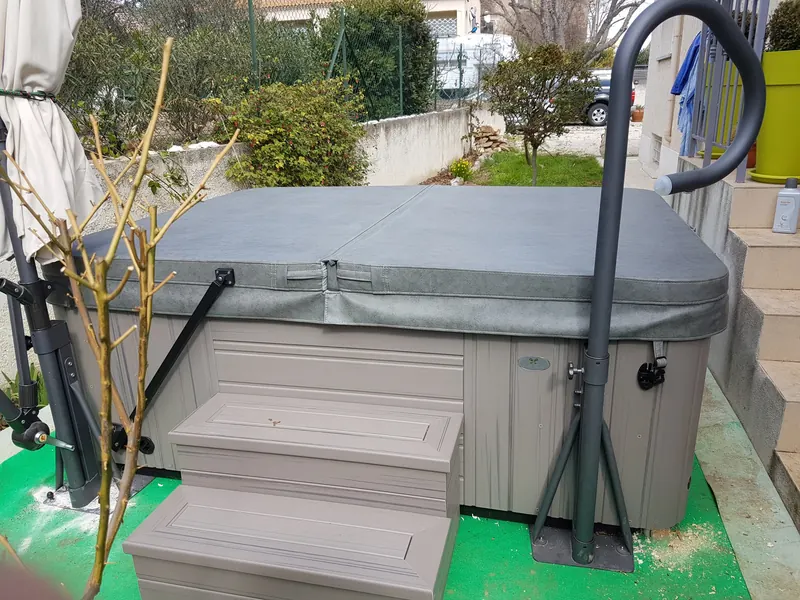 Spa sous sa couverture grise, avec marchepied, main courante et lève-couverture, posé sur un tapis vert dans un jardin