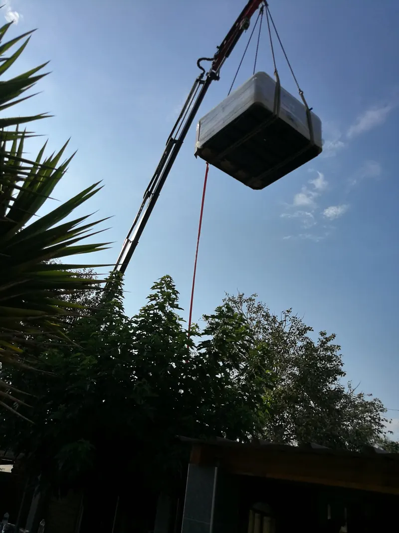 Spa emballé suspendu au bras d'une grue au-dessus des arbres et du toit d'un abri de jardin