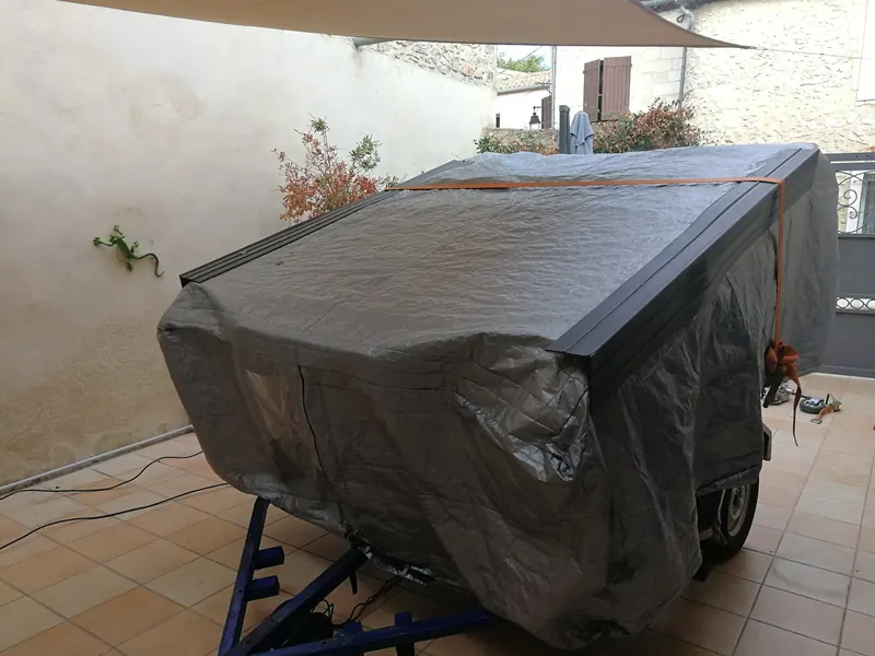 Spa emballé dans une bâche grise et sanglé sur une remorque, dans une cour carrelée sous une voile d'ombrage