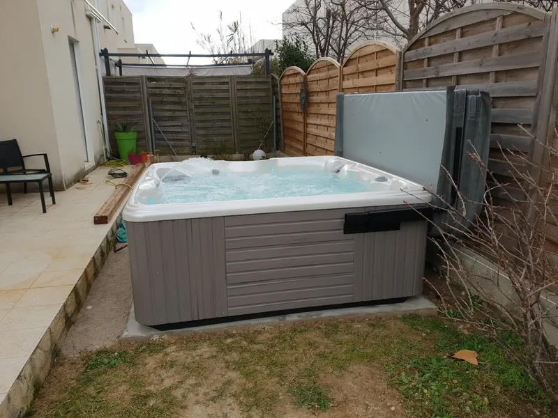 Spa gris en marche sur dalle béton au pied d'une terrasse carrelée, couverture repliée sur son support