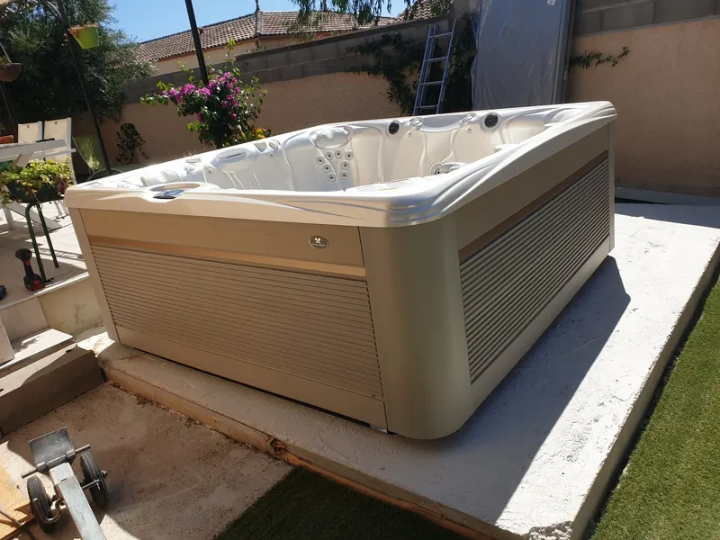 Spa Caldera blanc à habillage beige sur une dalle béton dans un jardin, chariot de transport au premier plan