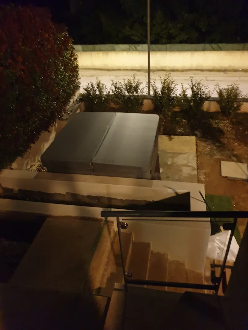 Spa fermé par une couverture grise dans un jardin, vu de nuit depuis un escalier à garde-corps vitré