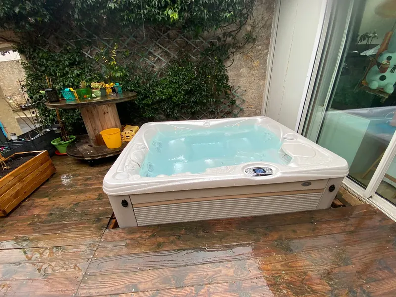 Spa blanc rempli d'eau turquoise sur la terrasse en bois, devant la baie vitrée