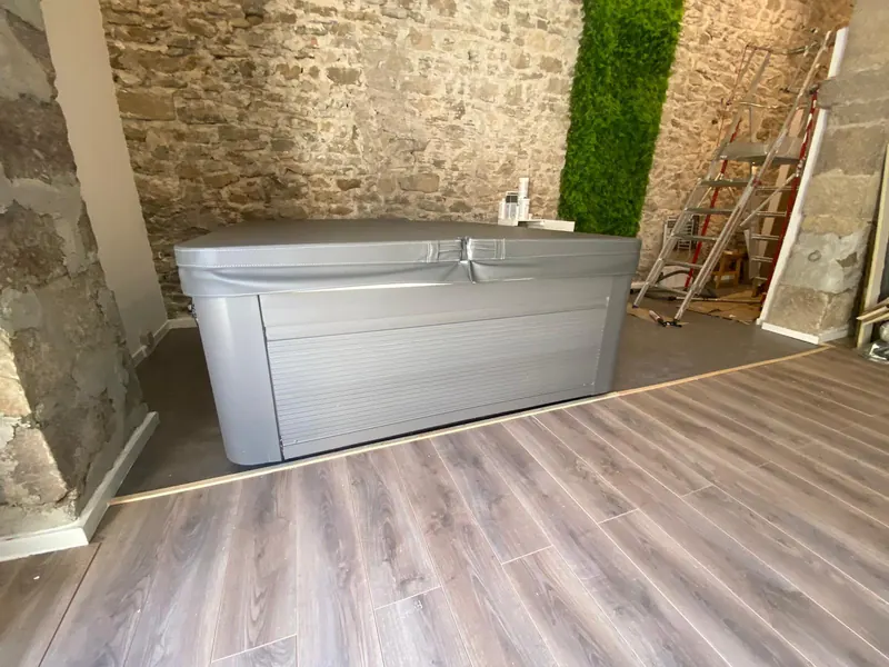Spa fermé sous son couvercle gris, mur de pierre, panneau végétal et escabeau derrière