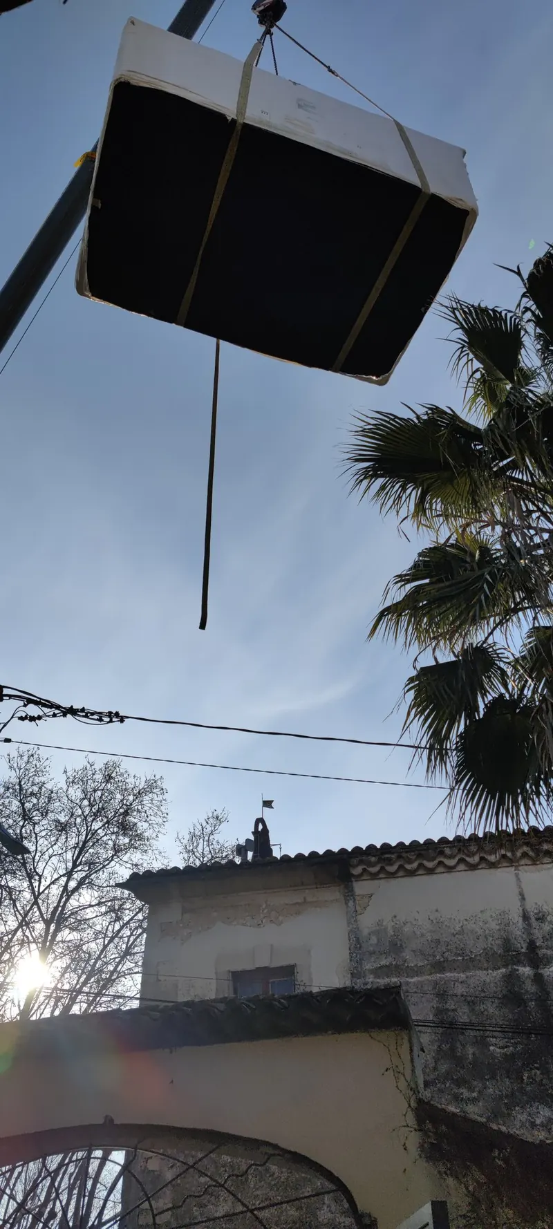 Spa emballé suspendu à la grue, vu d'en dessous, palmier et toits de tuiles en arrière-plan