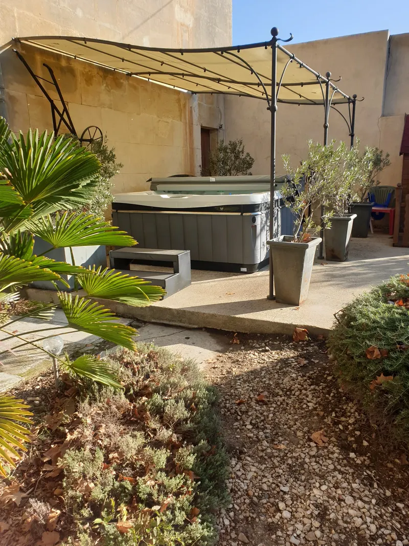 Spa gris installé sous une pergola, contre un mur en pierre, vu depuis le jardin