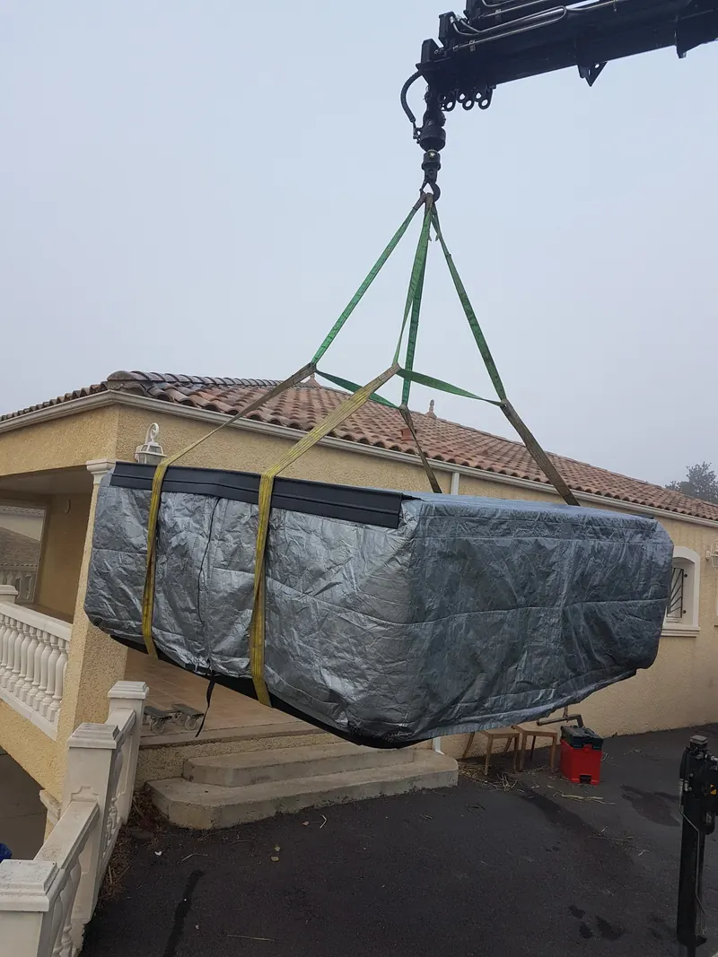 Bras de grue soulevant le spa bâché au-dessus de l'allée, devant la maison à Lespignan