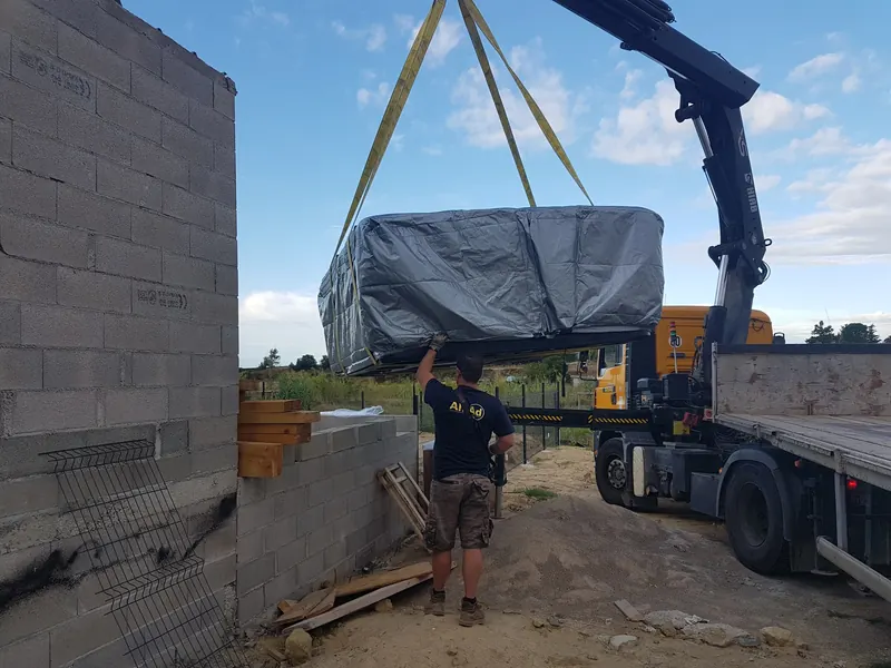 Spa sous bâche suspendu à la grue au-dessus d'un mur en parpaings, guidé à la main, à Mèze