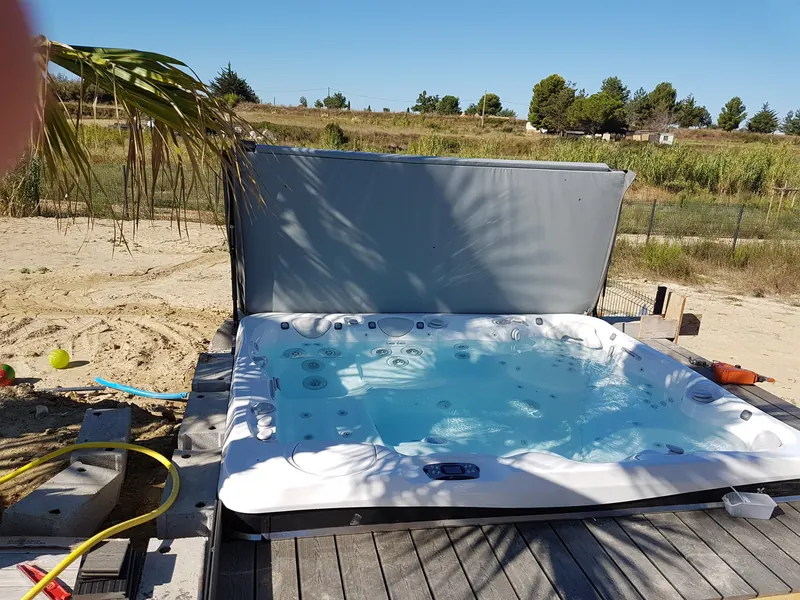 Spa rempli sur une terrasse en bois, couvercle relevé, à l'ombre d'un palmier à Mèze