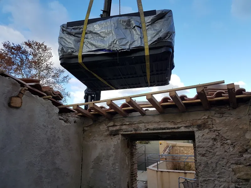 Spa emballé et sanglé, suspendu à la grue au-dessus d'une toiture en partie découverte à Montpeyroux