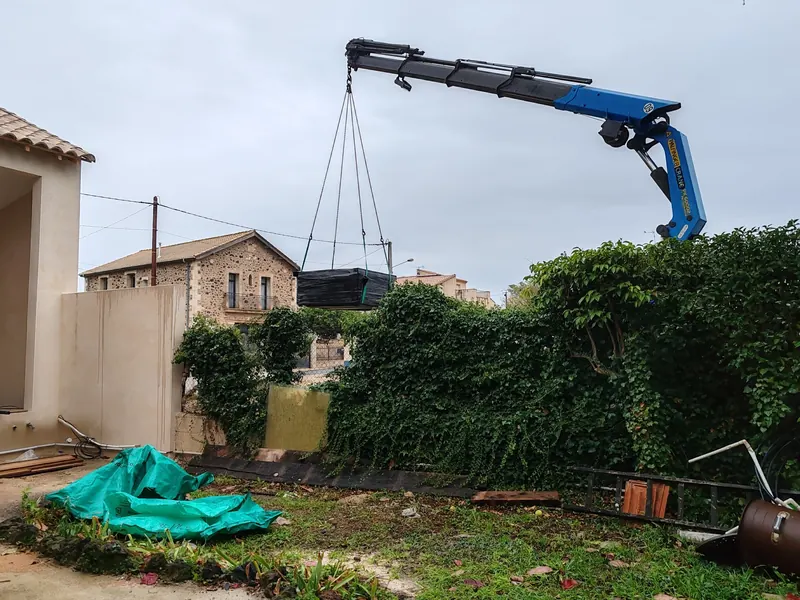 Spa emballé de film noir suspendu à la grue bleue au-dessus d'une haie, maison en pierre derrière