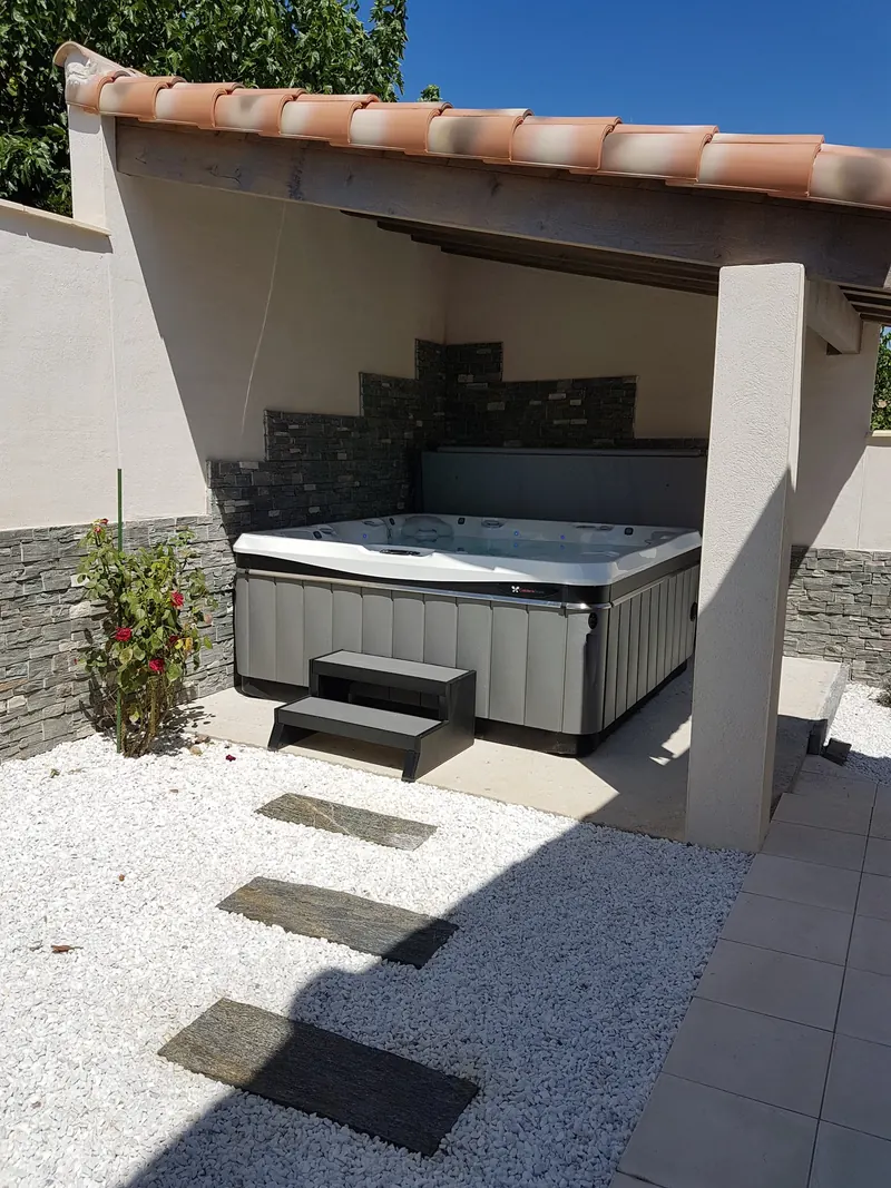 Spa à jupe grise et son marchepied sous l'abri, devant un mur en parement de pierre