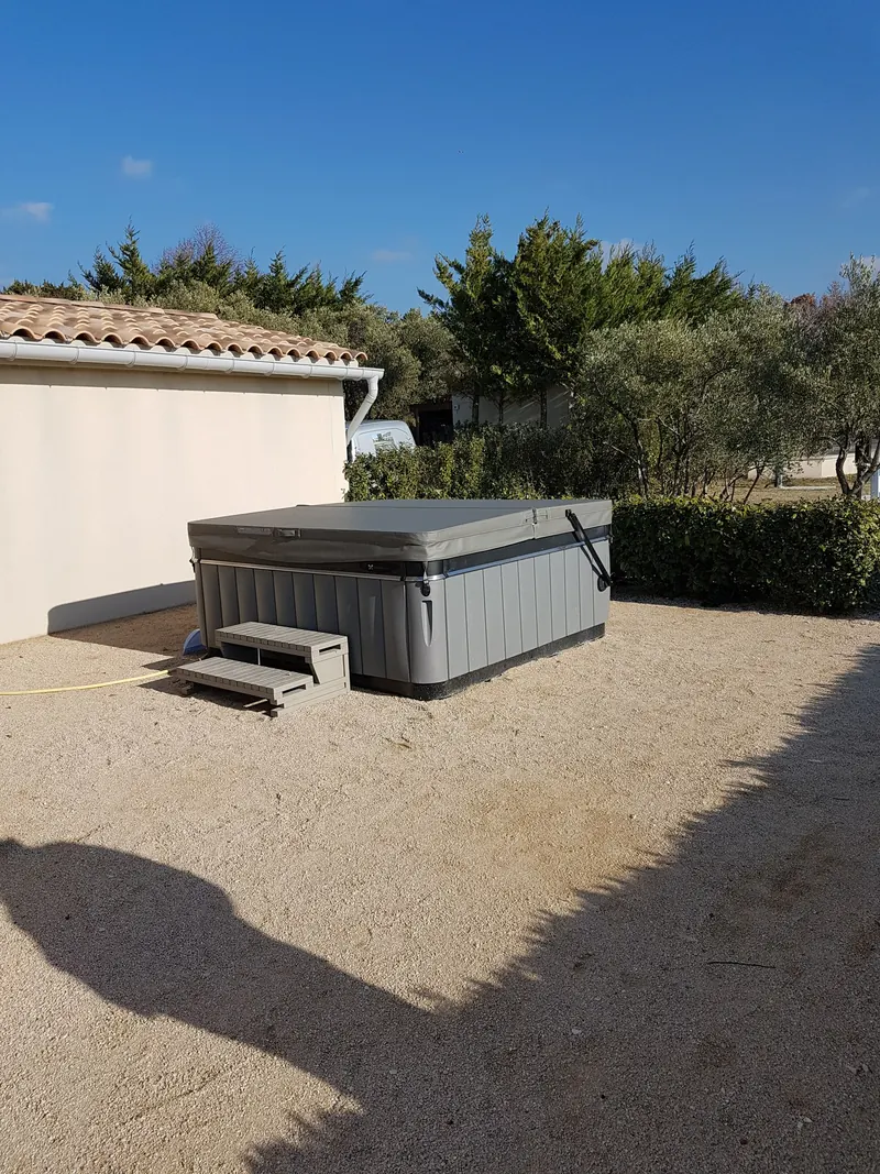 Spa gris couvert posé sur le gravier au pied d'une maison, avec son marchepied, à Saint-Pierre-de-Vassols