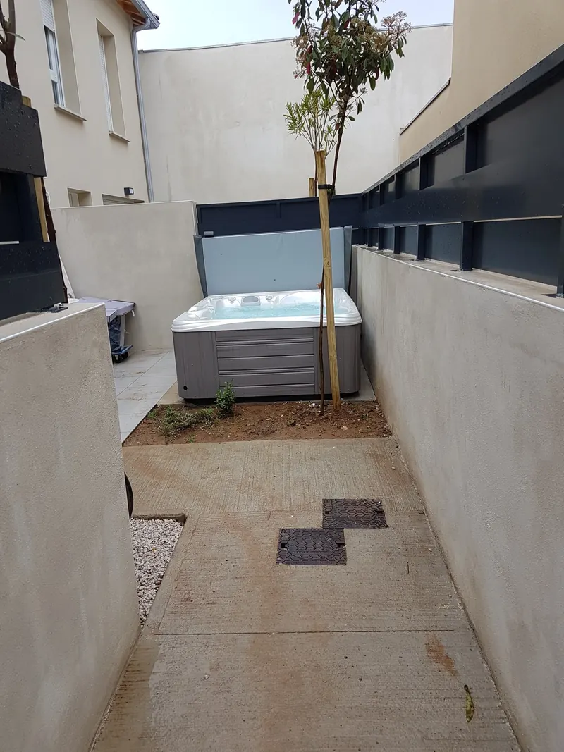 Allée bétonnée entre deux murs menant au spa gris posé au fond de la cour
