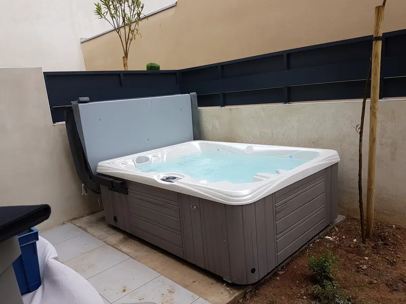 Spa en eau contre un muret et une clôture sombre, couverture relevée à l'arrière du spa