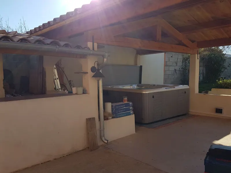 Spa installé sous la charpente d'une pergola, vu de côté depuis la terrasse, à contre-jour