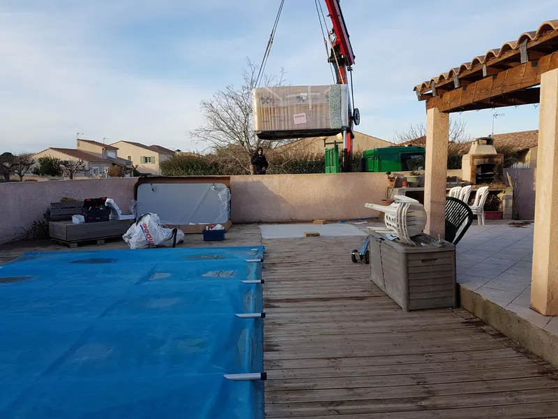 Spa emballé levé à la grue au-dessus du mur d'une terrasse en bois avec piscine bâchée à Loupian