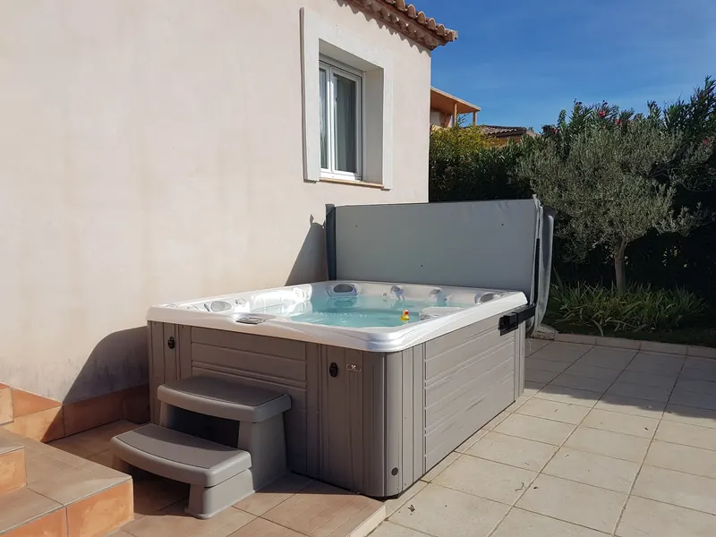 Spa installé dans l'angle d'une terrasse carrelée, avec marchepied, couvercle relevé et olivier