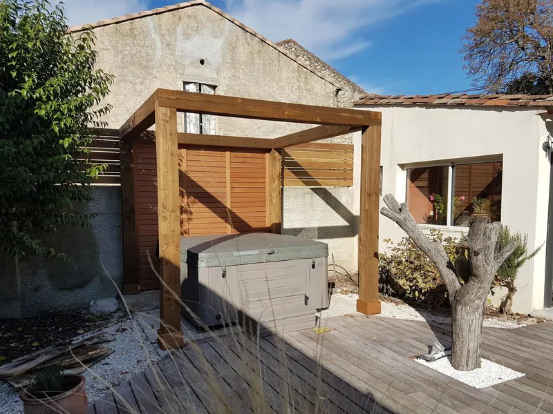 Pergola en bois abritant le spa, couvercle fermé, au bout de la terrasse devant la maison