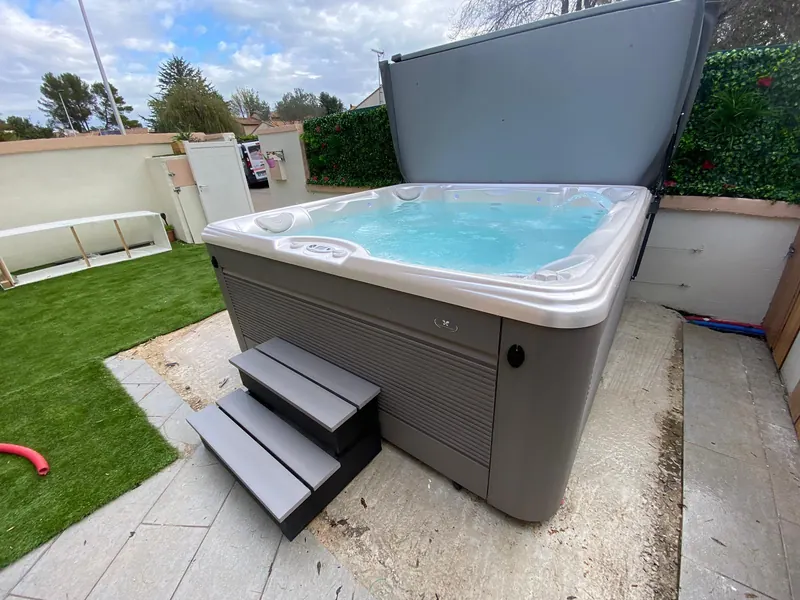 Spa Palatino blanc et gris installé dans le jardin, avec gazon, haie fleurie et abri