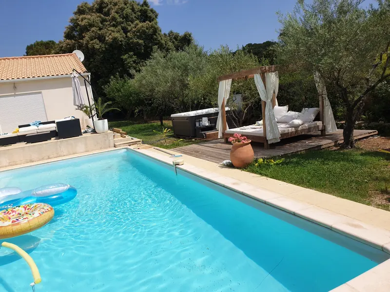 Piscine au premier plan, spa installé derrière, à côté d'un lit de jardin à baldaquin sur terrasse bois