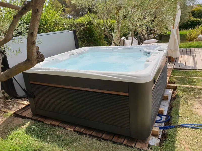 Spa rempli sous un olivier, couvercle relevé à l'arrière, sur son socle en bois