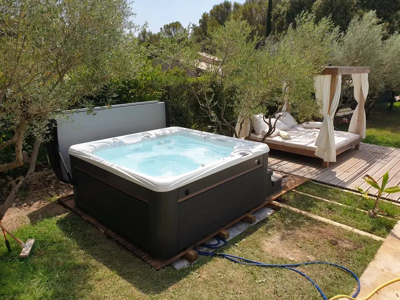 Spa en eau sur son socle en bois entre les oliviers, à côté d'un lit de jardin à baldaquin