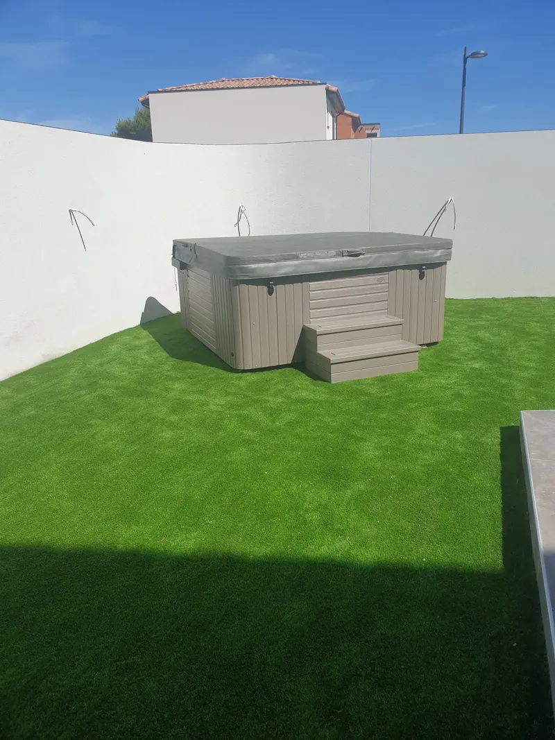 Spa taupe avec marchepied et couverture fermée, posé sur gazon synthétique contre un mur blanc à Vic-la-Gardiole