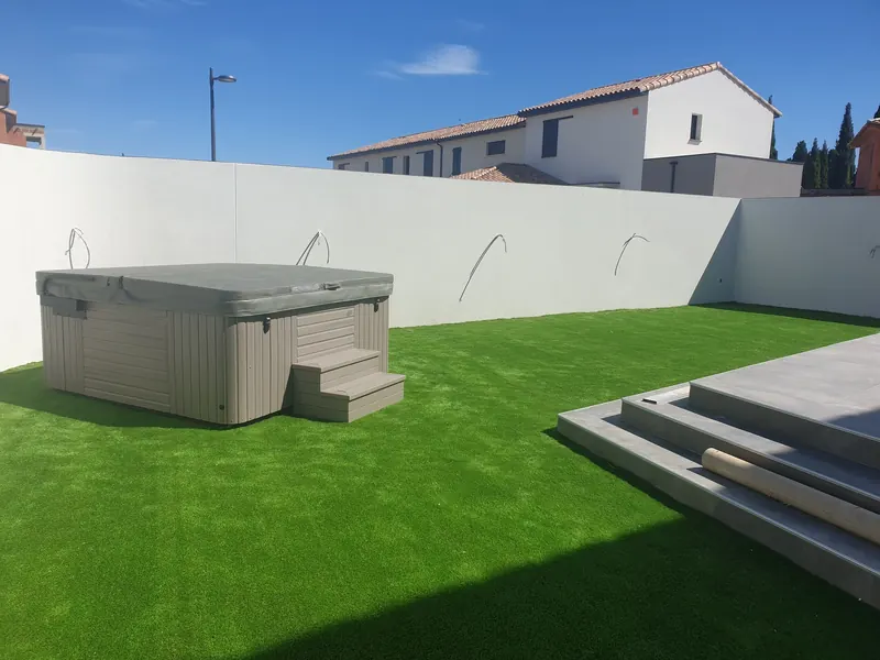 Vue large du jardin clos : spa couvert dans l'angle, gazon synthétique et marches de terrasse, à Vic-la-Gardiole