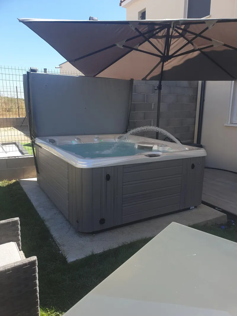 Spa gris sous un grand parasol, entre la maison et un mur en parpaings, à Cournonterral