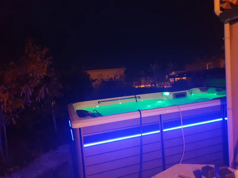 Spa de nage éclairé de nuit, eau verte et bandeau lumineux bleu sur l'habillage, à Bollène