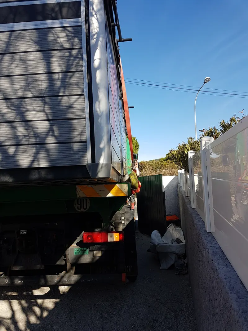 Camion-grue chargé du spa de nage, engagé dans un passage étroit entre la maison et la clôture