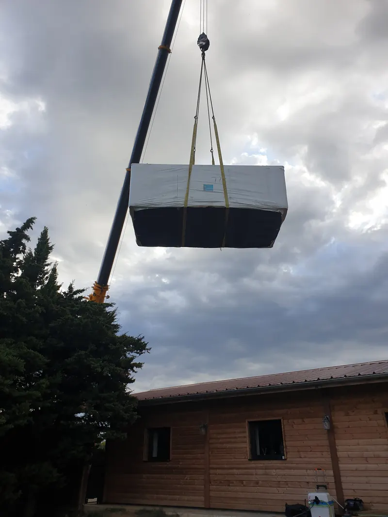Spa de nage emballé vu de dessous, suspendu à la flèche de la grue au-dessus d'une maison en bois