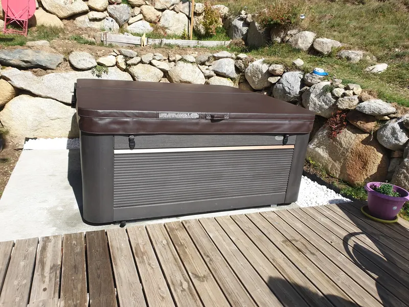 Spa gris à couvercle marron posé sur une dalle béton au pied d'un mur de pierres à Égat