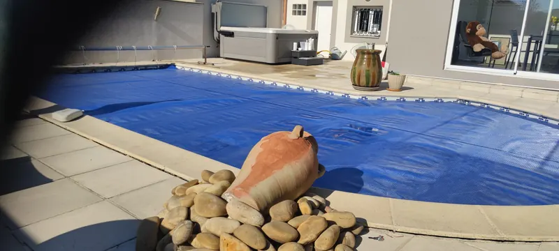 Piscine à bâche bleue et amphore au premier plan, spa visible derrière sur la terrasse