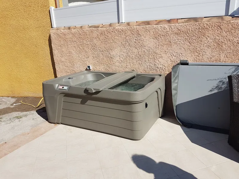 Spa Fantasy Spas Aspire gris taupe rempli d'eau, posé sur une terrasse carrelée contre un mur crépi à Mèze