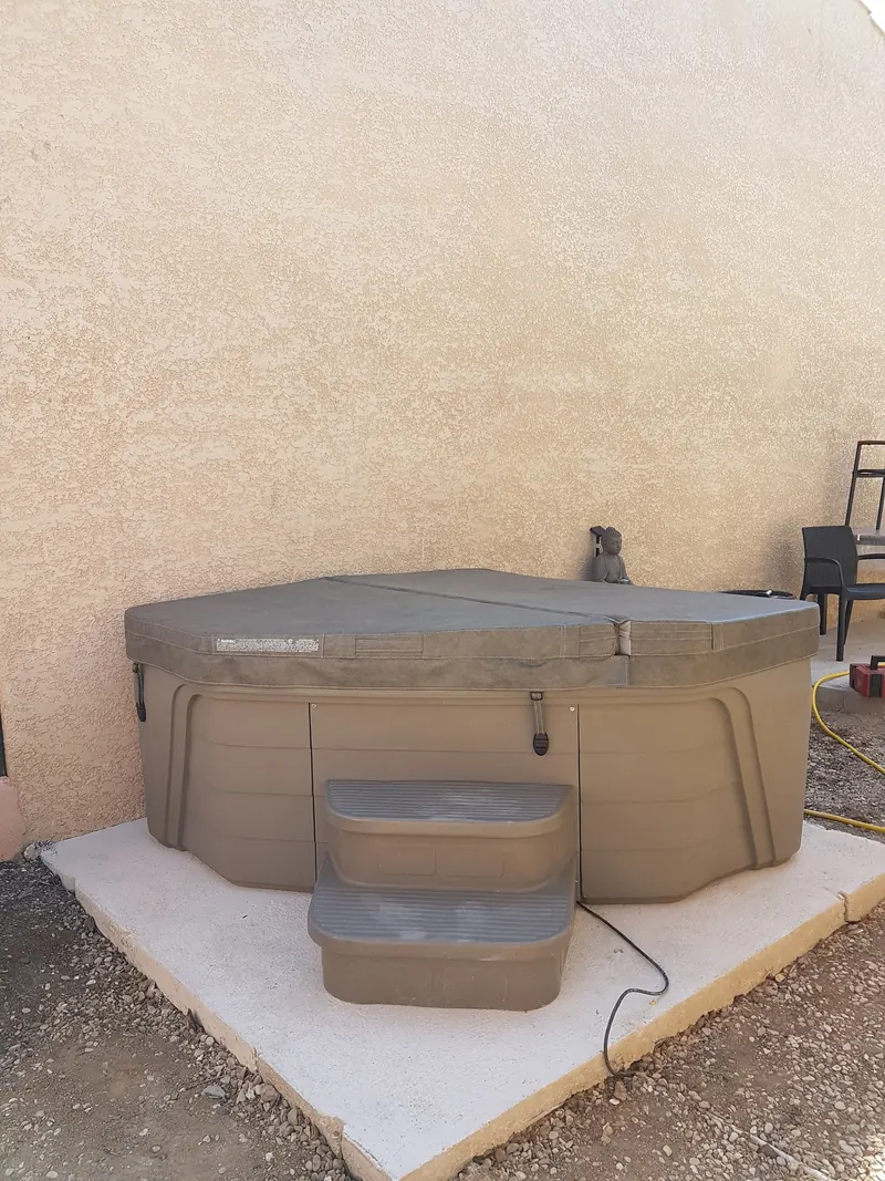 Spa taupe fermé par son couvercle gris, posé sur une dalle béton contre un mur crépi à Mèze