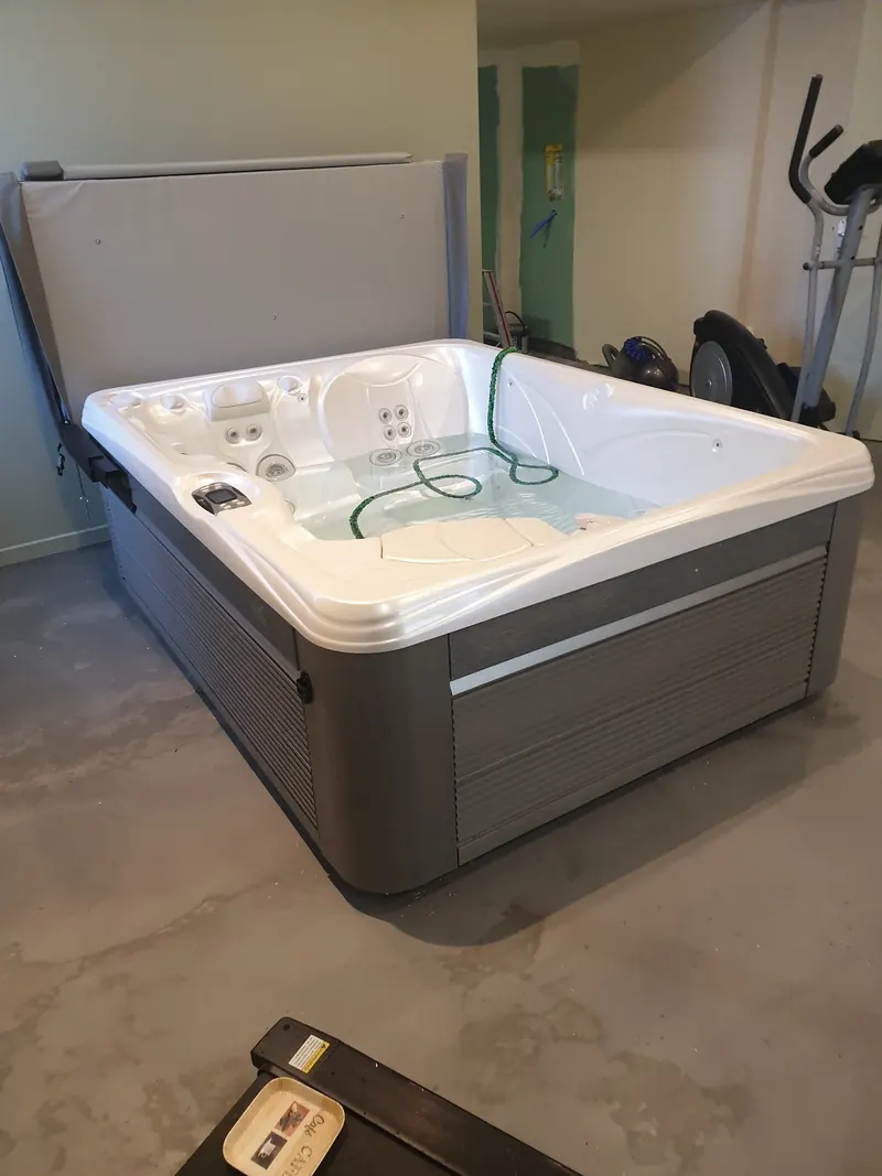 Spa blanc et gris en cours de remplissage au tuyau, couverture relevée, dans une pièce intérieure à Gigean